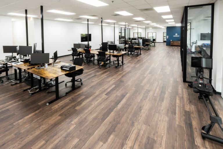 A modern office for Echelon with wooden floors includes desks with computers and ergonomic chairs, illuminated by fluorescent lights and large windows. An advanced rowing machine near a sleek glass partition adds a fitness touch.