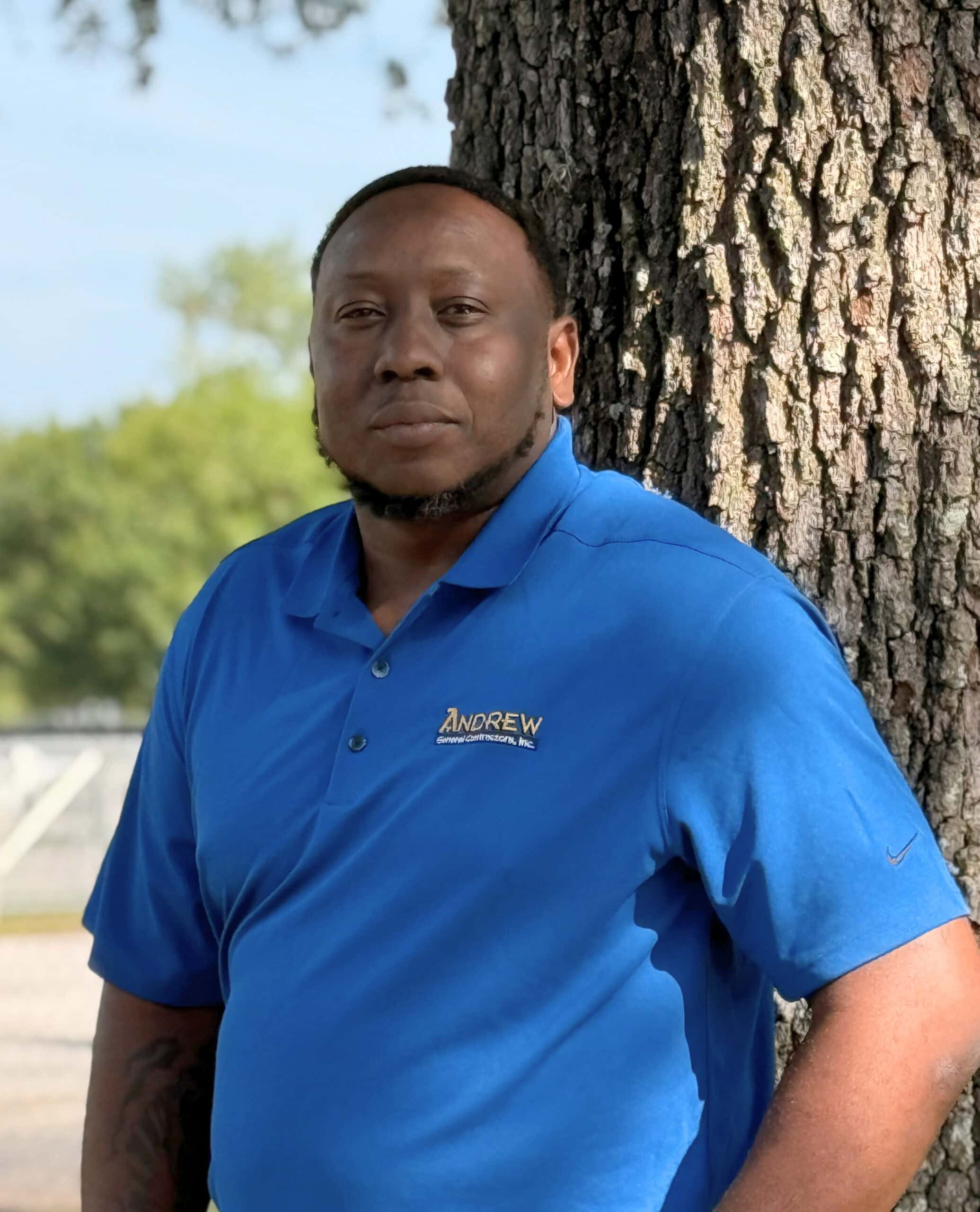 A man in a blue ANDREW company polo representing an Orlando commercial general contractor leans against a tree outdoors, with greenery and a fence in the background.