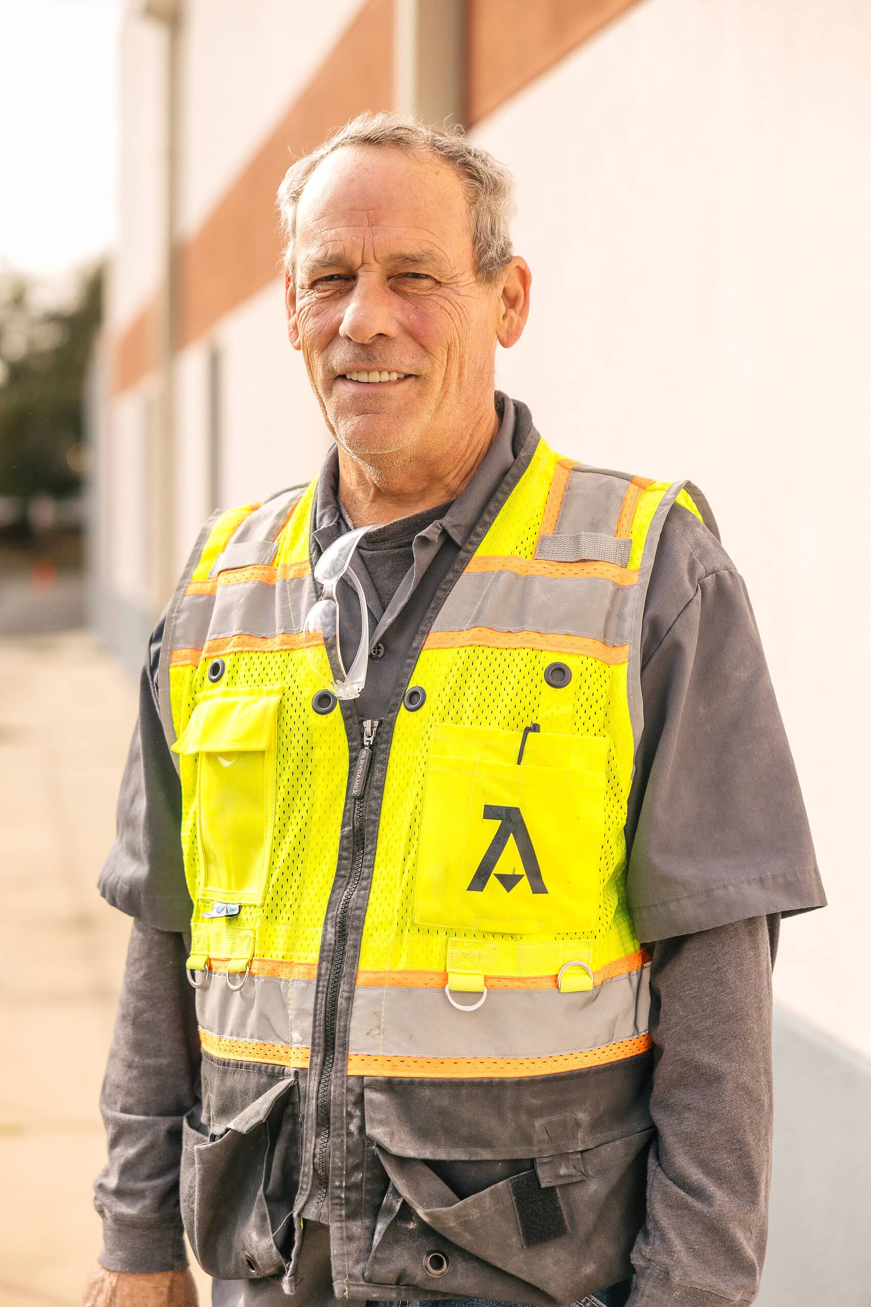 Older man in a gray shirt and yellow safety vest stands outside a building in Orlando, smiling slightly at the camera as a Commercial General Contractor.