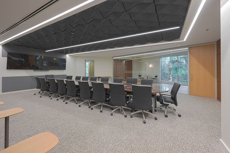 Modern conference room with an oval table, sixteen black chairs, a wall-mounted screen, and floor-to-ceiling windows facing greenery.